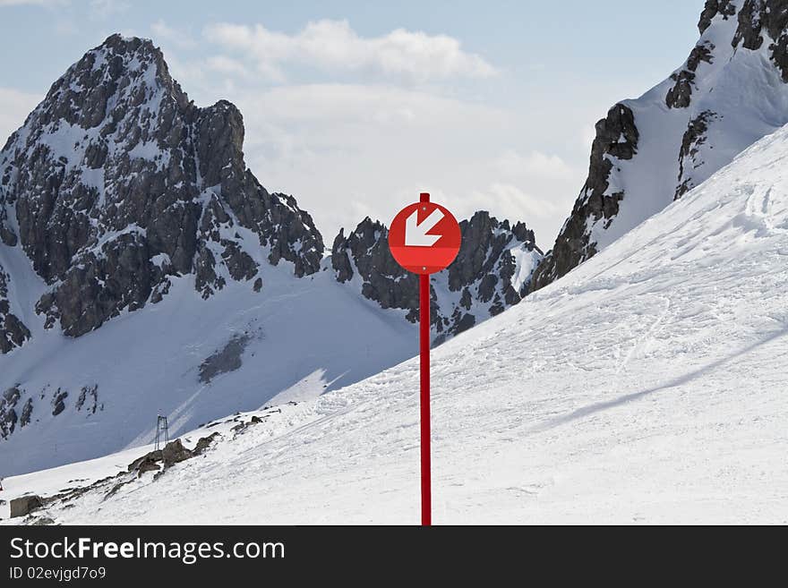 Red sign with arrow pointing down showing direction of ski run with snowy mountain peaks behind. Red sign with arrow pointing down showing direction of ski run with snowy mountain peaks behind