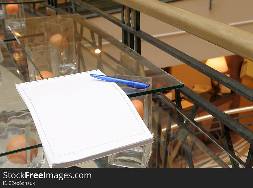 Blank writing pad with a pen lying on a glass table at the hotel lobby. Blank writing pad with a pen lying on a glass table at the hotel lobby.