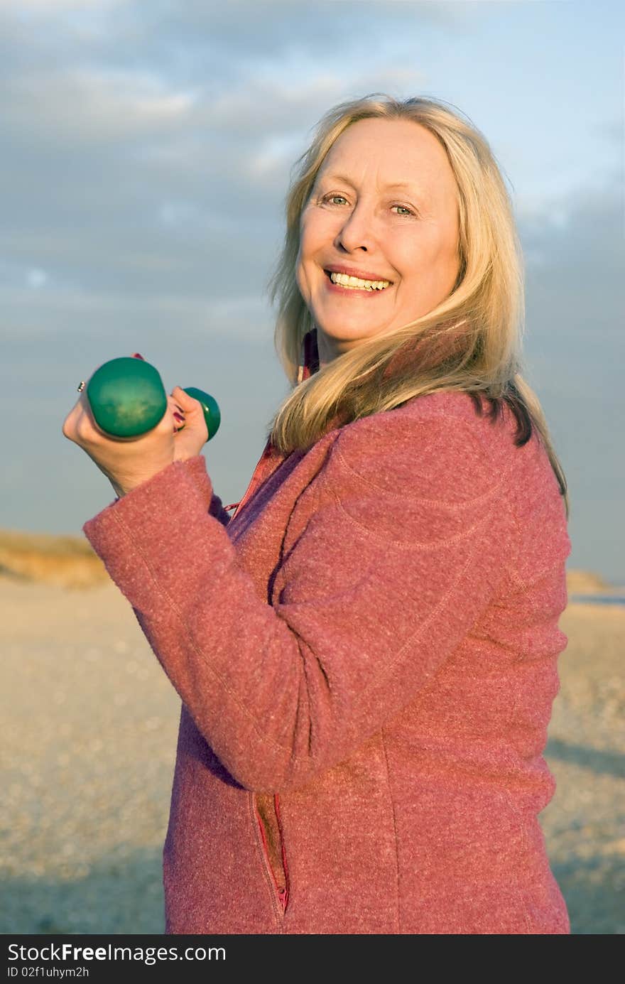 A colour portrait photo of a beautiful happy smiling older lady in her sixties exercising with dumbells on the beach. A colour portrait photo of a beautiful happy smiling older lady in her sixties exercising with dumbells on the beach.