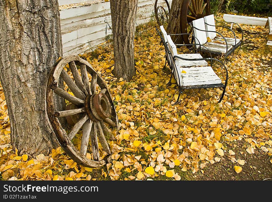 Old weathered wooden wagon wheel leaning against tree trunk with benches in BG and yellow leaves on groound. Old weathered wooden wagon wheel leaning against tree trunk with benches in BG and yellow leaves on groound.