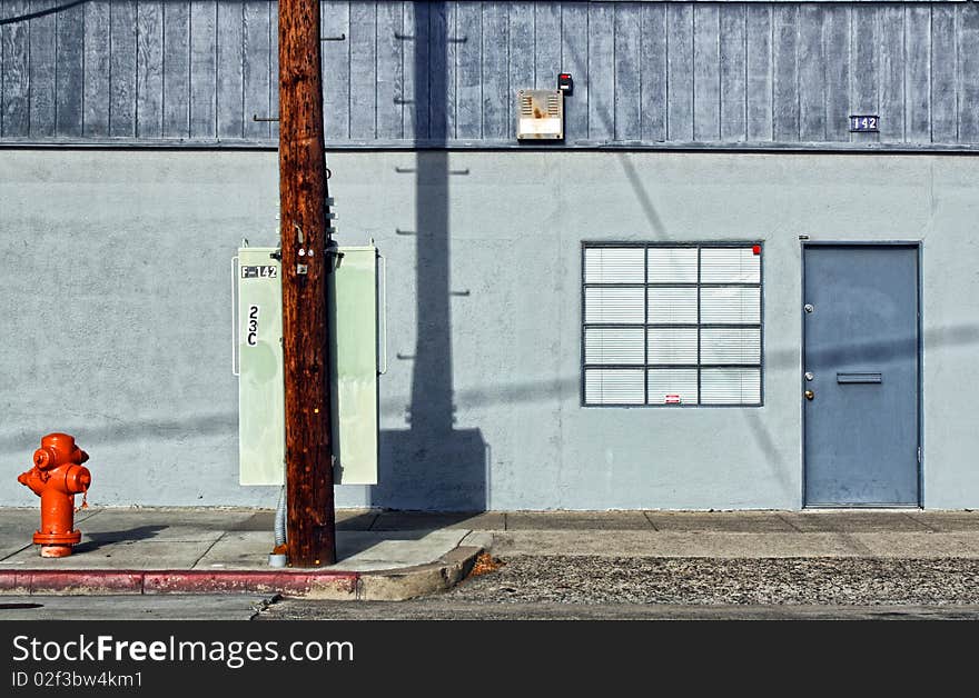 The side door of a gray, industrial building is seen along an urban sidewalk, with a bright orange fire hydrant and telephone pole in the foreground. The side door of a gray, industrial building is seen along an urban sidewalk, with a bright orange fire hydrant and telephone pole in the foreground.