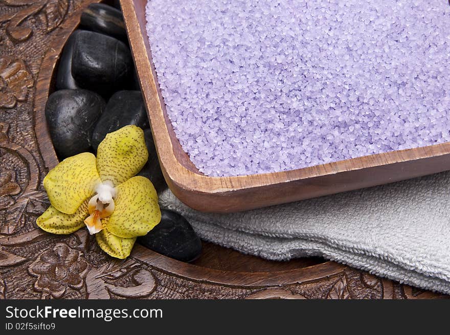 Spa scene with bath salts, massage stones, towel and an orchid on in a carved wood setting.