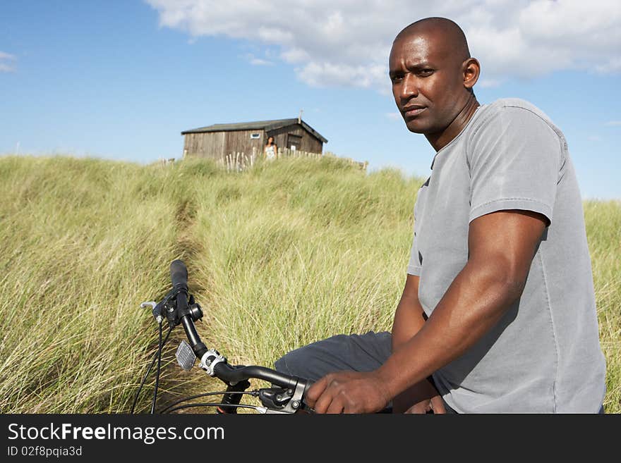 Young Man Riding Mountain Bike By Dunes With Old Beach Hut In Distance