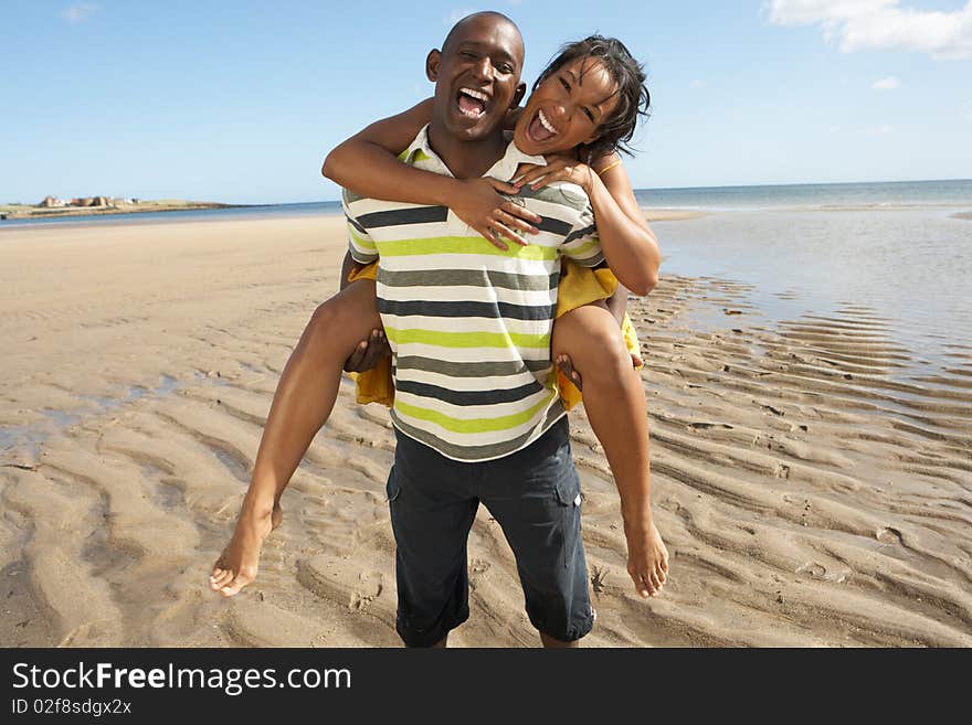 Young Man Giving Woman Piggyback Along Shoreline Of Beach