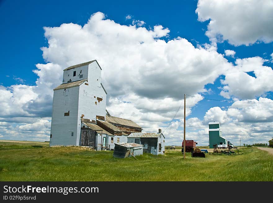 Olds weathered grey grain elevator against blue sky with clouds, green grass in foreground and dark green grain elevator in distance. Olds weathered grey grain elevator against blue sky with clouds, green grass in foreground and dark green grain elevator in distance.