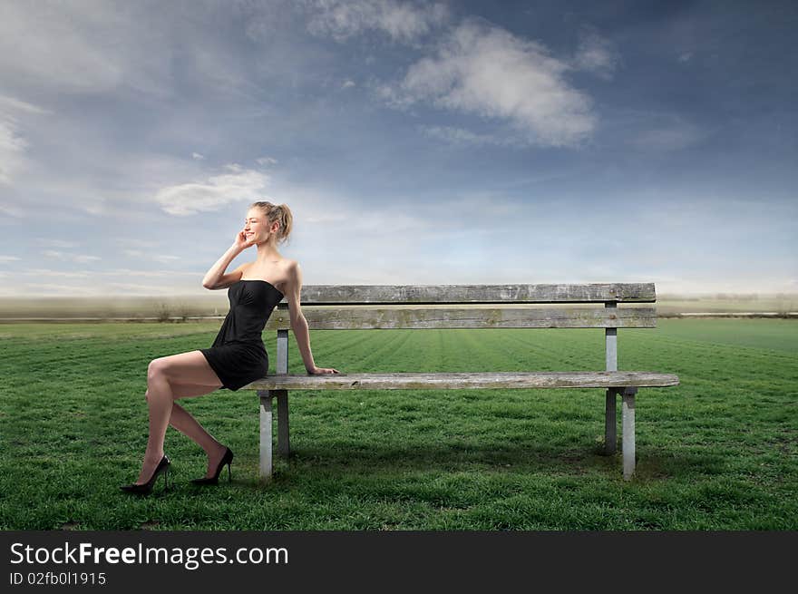 Elegant young woman using a mobile phone while sitting on a park bench on a green meadow. Elegant young woman using a mobile phone while sitting on a park bench on a green meadow