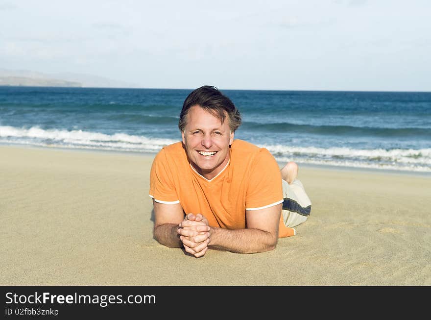 A colour portrait photo of a happy smiling forties man laying on a beautiful sandy beach.