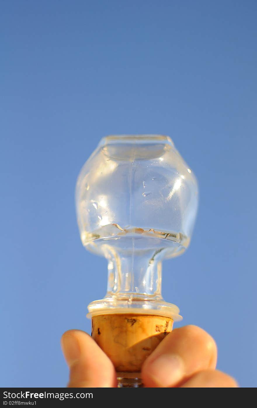 A glass stopper with a cork bottom held up against a clear blue morning sky reflecting glints of sunlight. A glass stopper with a cork bottom held up against a clear blue morning sky reflecting glints of sunlight.