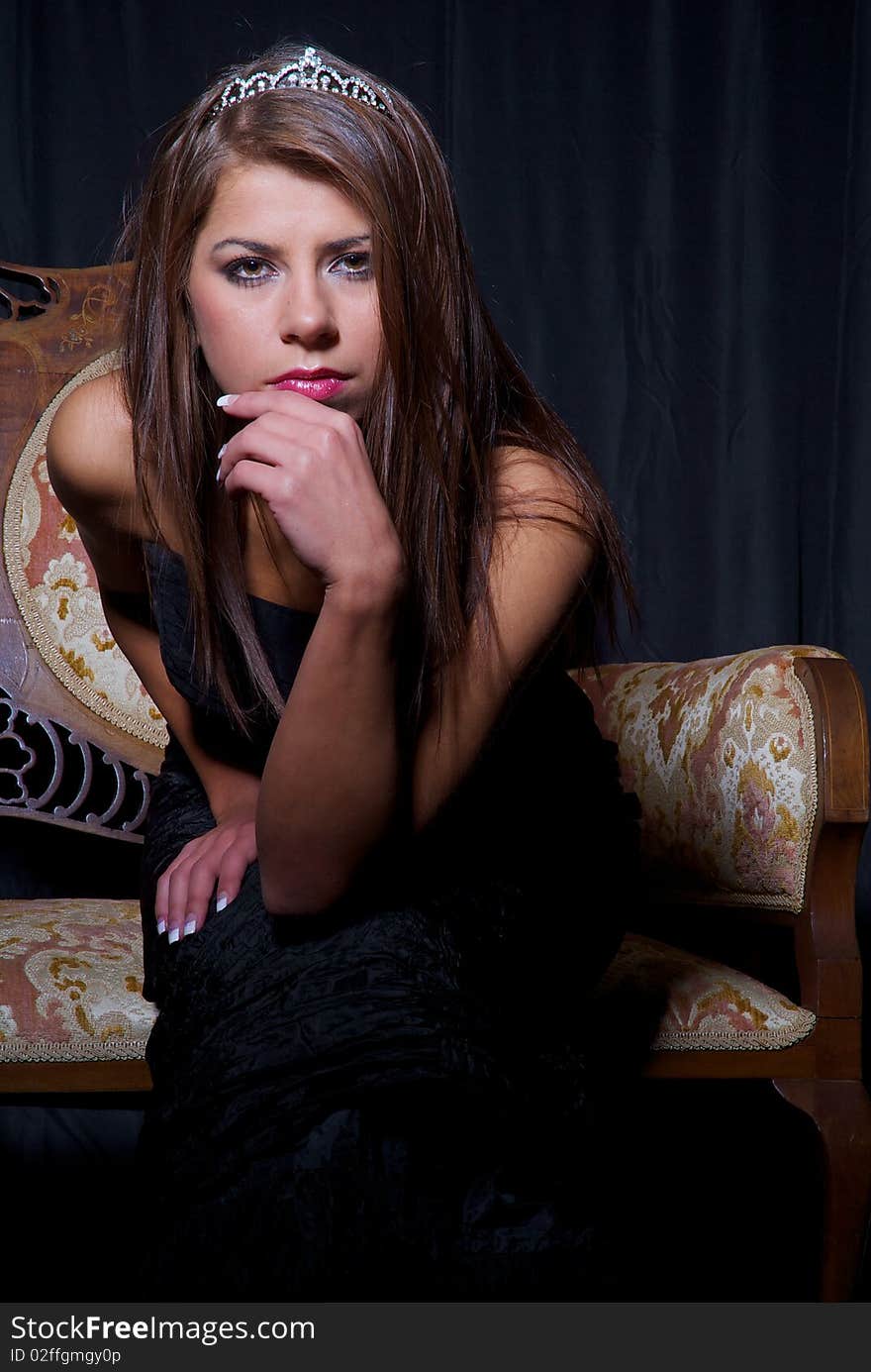 Girl wearing black prom dress and a crown sitting on the antique sofa, black background. Girl wearing black prom dress and a crown sitting on the antique sofa, black background