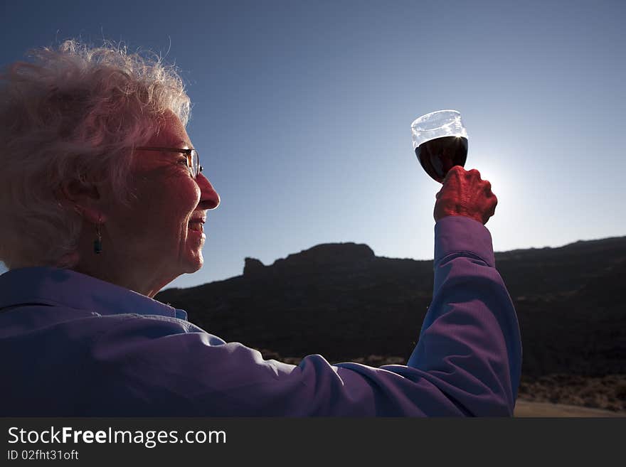 An elderly woman is in a desert landscape and holding up a glass of red wine in a toast. Horizontal shot. An elderly woman is in a desert landscape and holding up a glass of red wine in a toast. Horizontal shot.