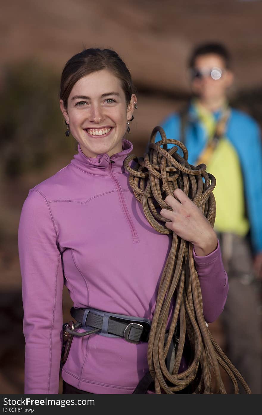 An attractive young woman is smiling and holding a climbing rope as her partner stands in the background. Vertical shot. An attractive young woman is smiling and holding a climbing rope as her partner stands in the background. Vertical shot.