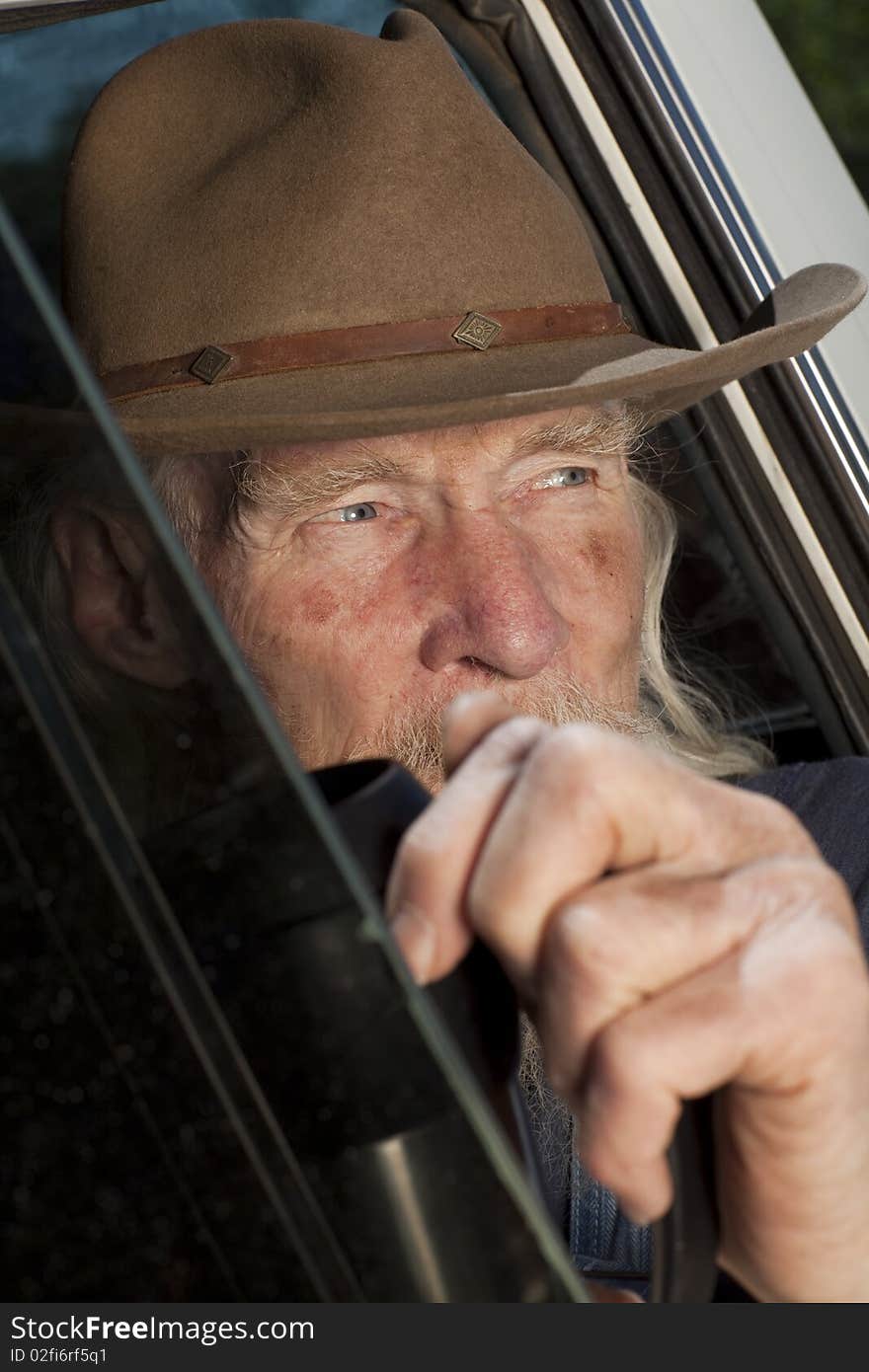 Cropped close-up of an elderly man with a cowboy hat and white beard, driving a pickup truck and staring out the window. Vertical format. Cropped close-up of an elderly man with a cowboy hat and white beard, driving a pickup truck and staring out the window. Vertical format.