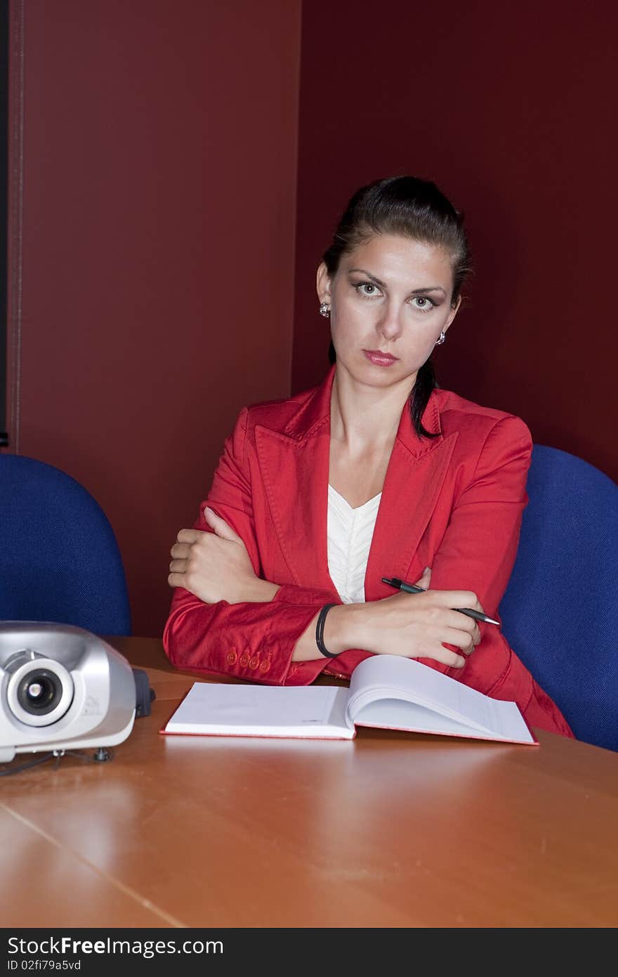 Attractive young businesswoman in a red blazer sits with her arms crossed and wears a serious expression on her face. Vertical shot.