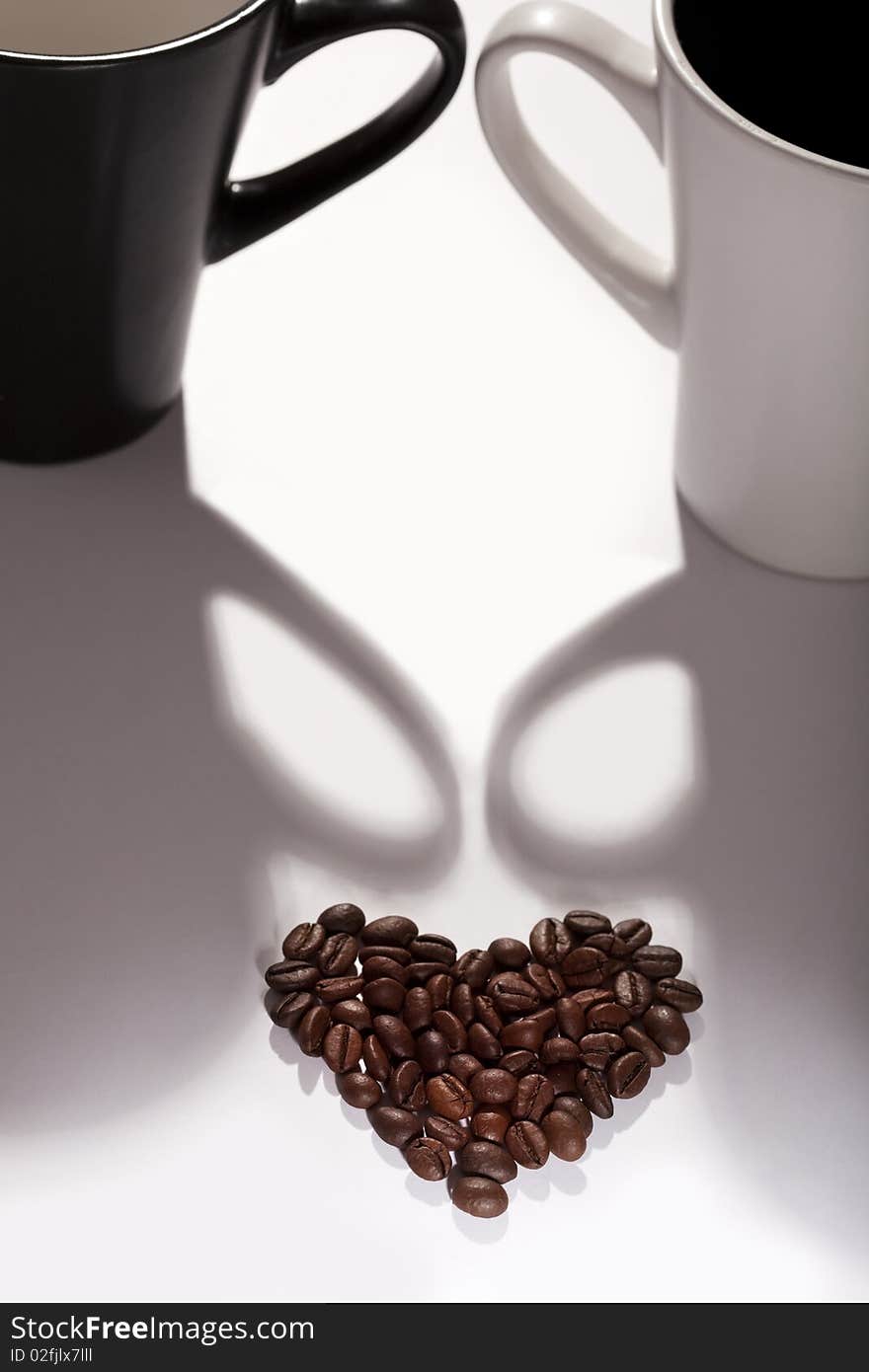 Various coffee beans in the shape of a heart against two cups, black and white