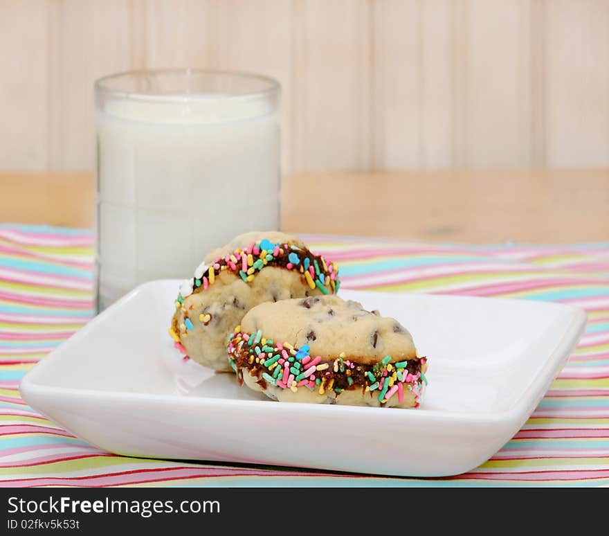 Plate with two chocolate chip cookie sandwiches with colorful sprinkles on a striped place mat with a glass of milk