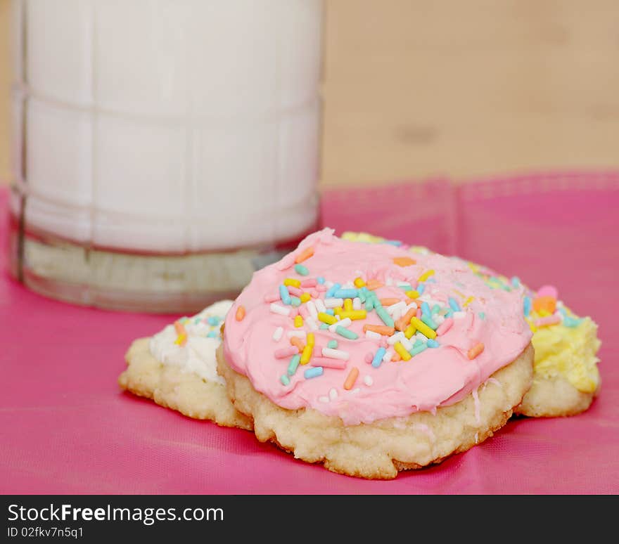 Three sugar cookies with icing and sprinkles with a glass of milk in the background on a pink place mat. Three sugar cookies with icing and sprinkles with a glass of milk in the background on a pink place mat