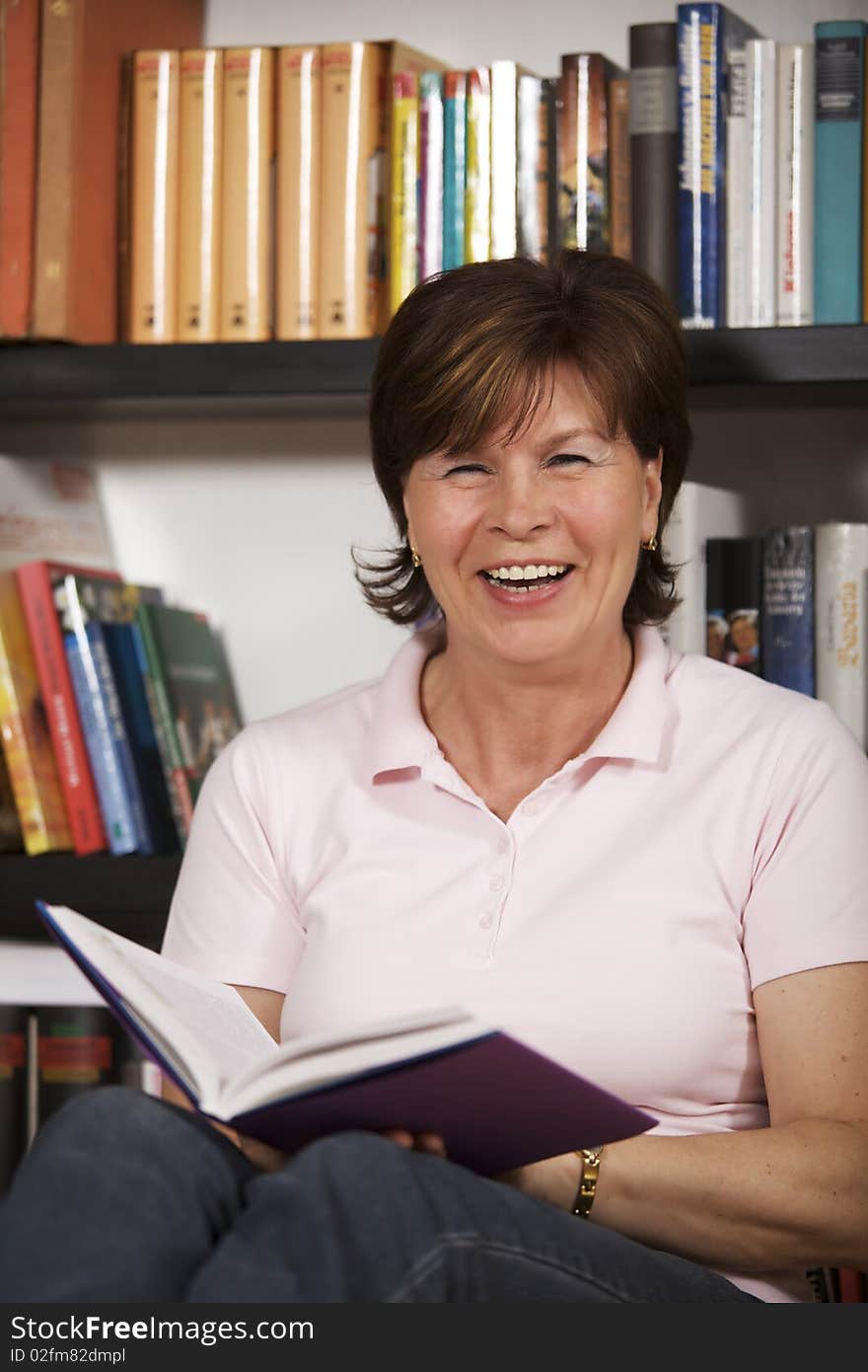 Laughing senior woman sitting on floor in front of bookshelf at home and reading a book. Laughing senior woman sitting on floor in front of bookshelf at home and reading a book.