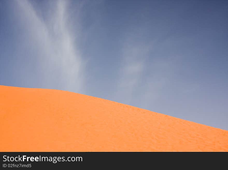 A featureless orange sand dune against a deep blue sky with whispy clouds