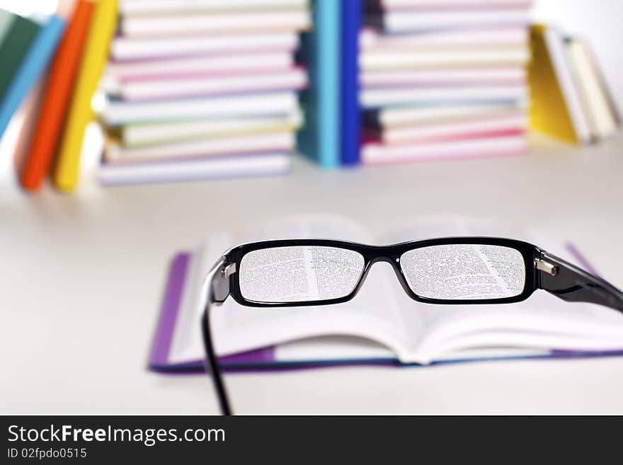 Clear view through specs facing open book in front of stack in rainbow colors paper wrapped books on white background, PHOTOGRAPH, NOT 3D RENDER.