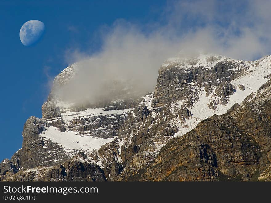 Snow covered mountain peak with clouds around it and the moon in the background