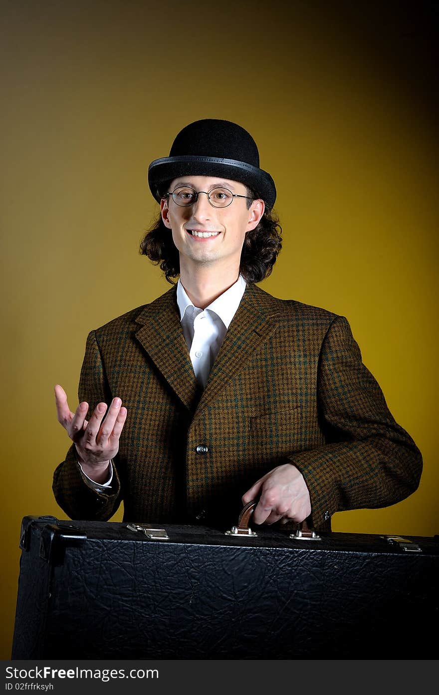 Portrait of young english gentleman in bowler hat holding suitcase as a luggage. yellow background