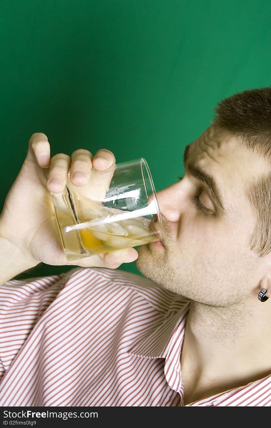 A young man dressed in a striped shirt on a green background makes the drink of whiskey on the rocks. A young man dressed in a striped shirt on a green background makes the drink of whiskey on the rocks