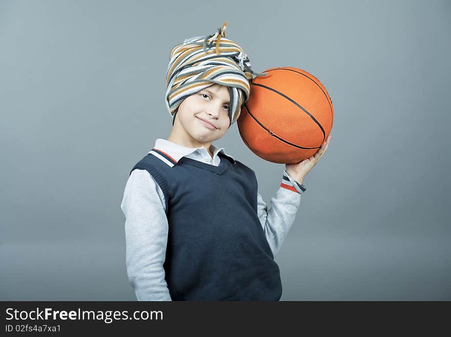 Young and funny caucasian tenager boy holding basketball ball near head and having fun with scarf around head standing isolated on gray background. Young and funny caucasian tenager boy holding basketball ball near head and having fun with scarf around head standing isolated on gray background