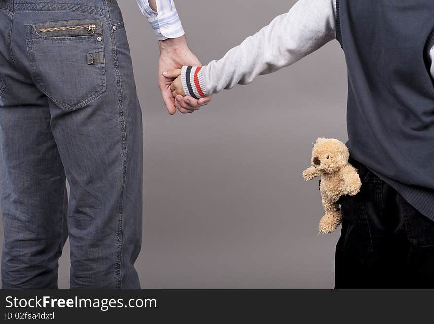 Young and careful father together with his teenager son holding hands of each other standing isolated over gray background with teddy bear. Young and careful father together with his teenager son holding hands of each other standing isolated over gray background with teddy bear