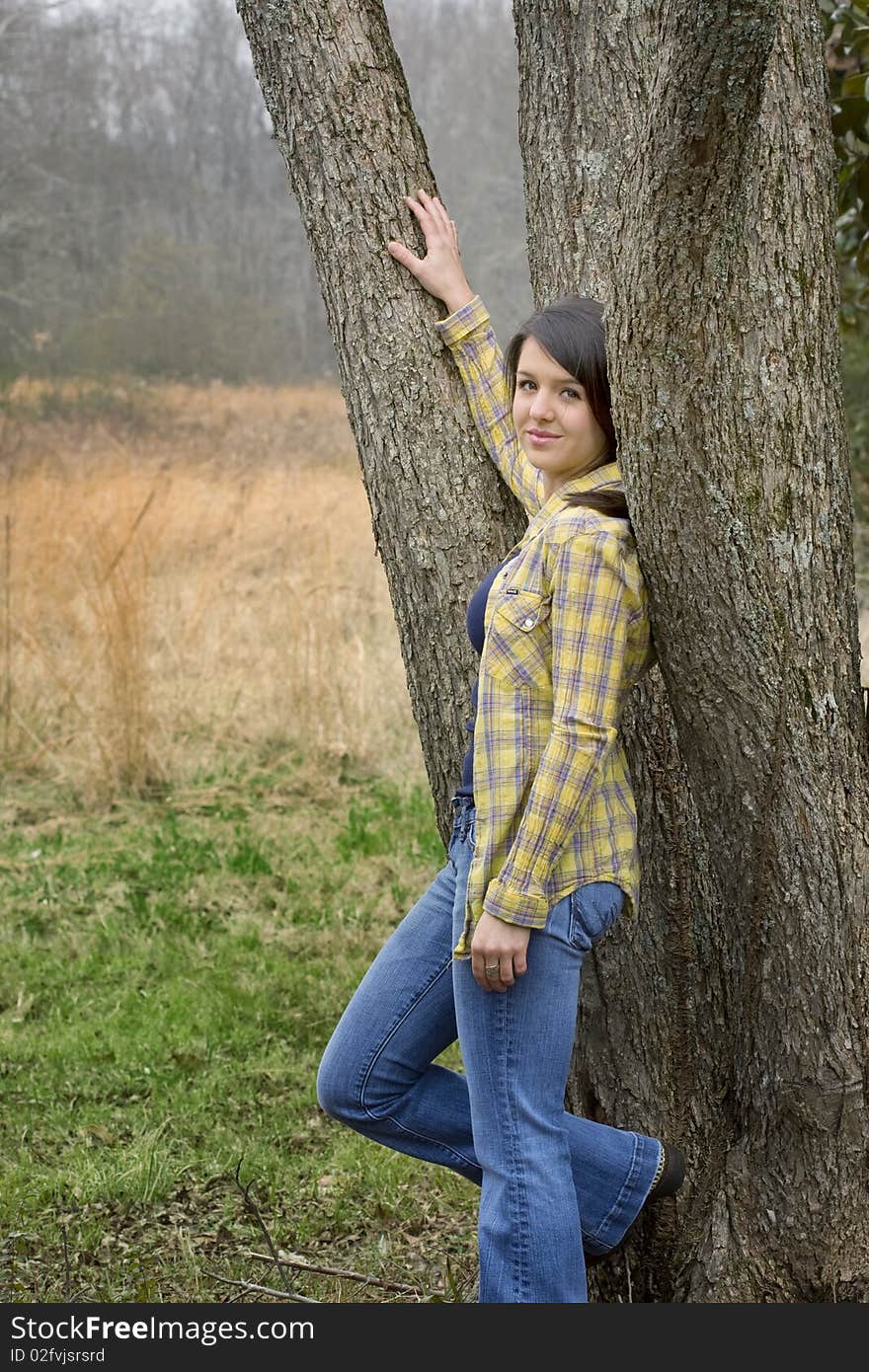 Young woman in yellow shirt and jean leaning against tree in pasture. Young woman in yellow shirt and jean leaning against tree in pasture