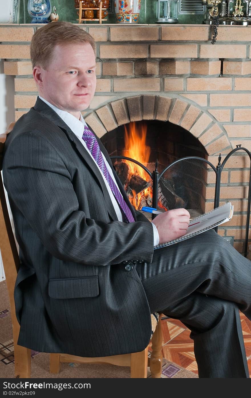 The man in a grey suit with a ball-point pen and a notebook at a house fireplace. The man in a grey suit with a ball-point pen and a notebook at a house fireplace.