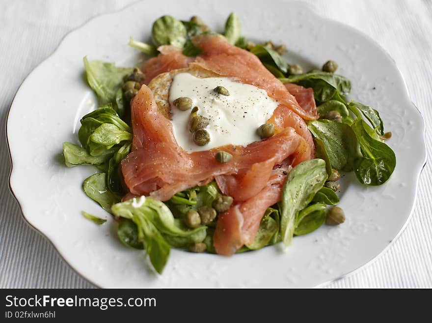 Smoked salmon salad with blinis and capers with sour cream on a white rustic plate.