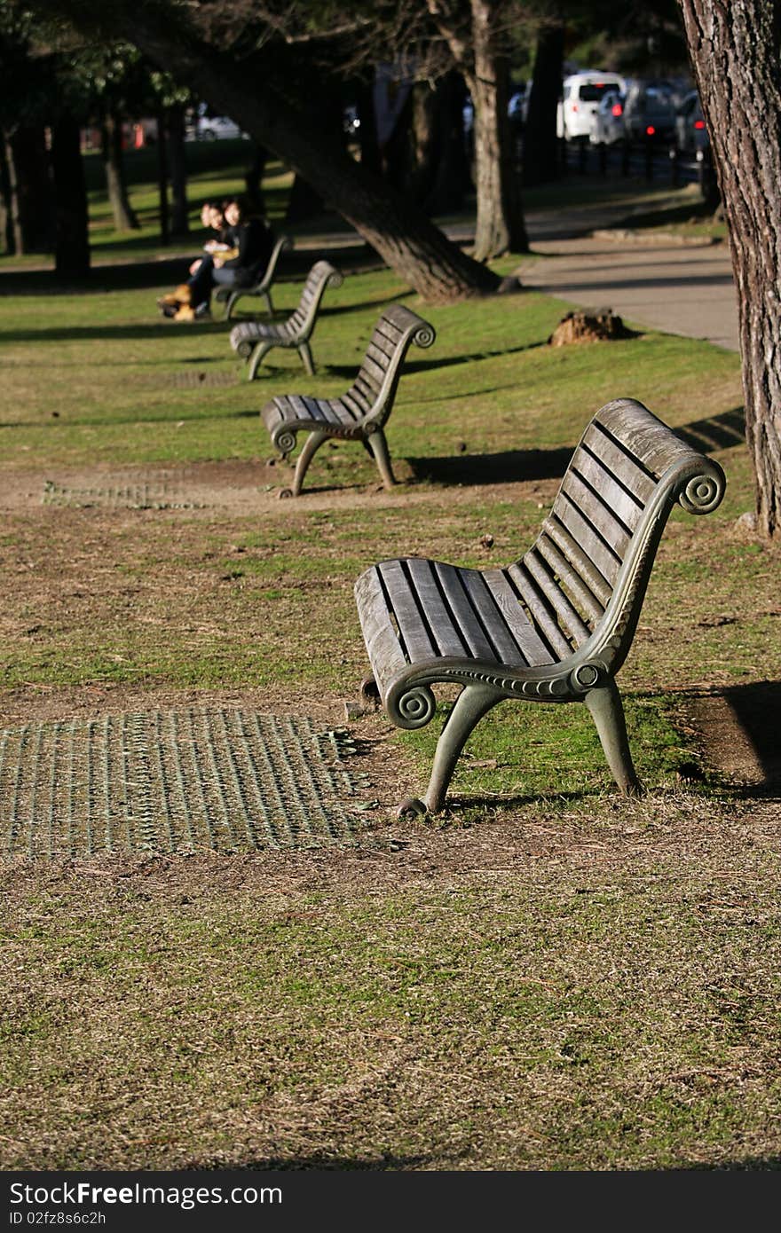 A row of benches bask in the sunshine, with people in the distance relaxing too. A row of benches bask in the sunshine, with people in the distance relaxing too.