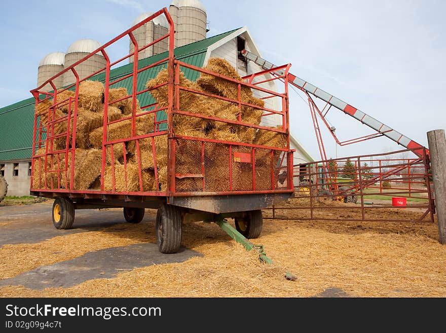 A photo of a hay wagon containing bales of straw. In the background is a barn with silos.