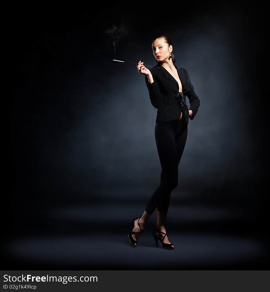 The portrait of a slim young lady in dark clothes holding a lighted cigarette in her hands. The portrait of a slim young lady in dark clothes holding a lighted cigarette in her hands