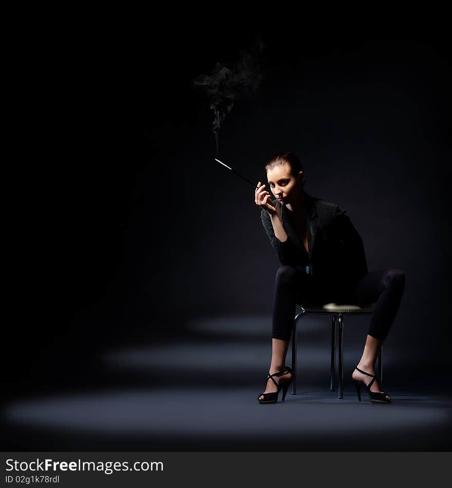 The portrait of a slim young lady in dark clothes holding a lighted cigarette in her hands. The portrait of a slim young lady in dark clothes holding a lighted cigarette in her hands