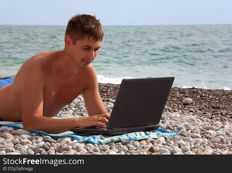 Young man working on his notebook on seacoast. portrait of guy with laptop lying on beach.