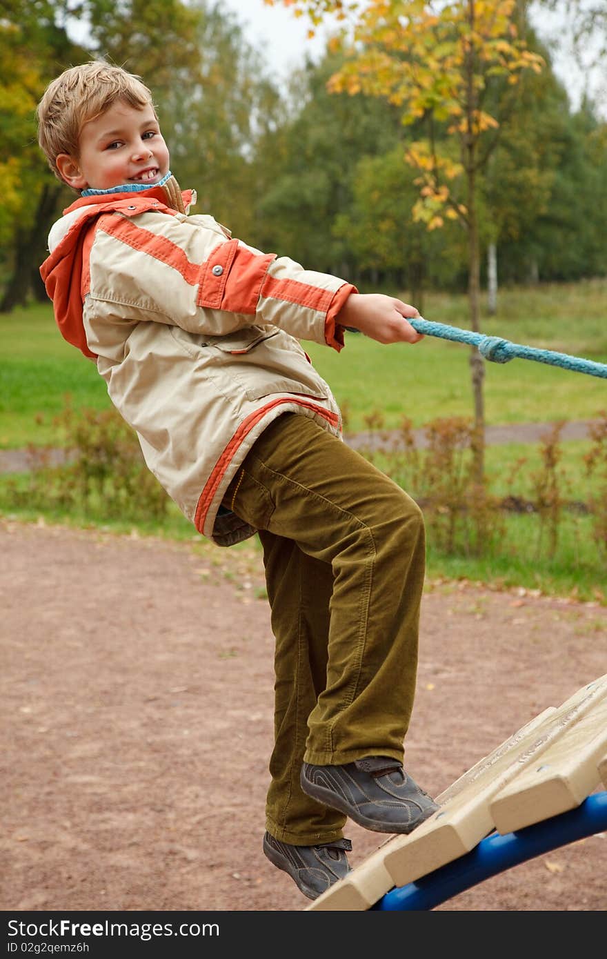 Boy in jacket is on playground in autumn park. Smiling, he climbs the stairs holding onto a rope.