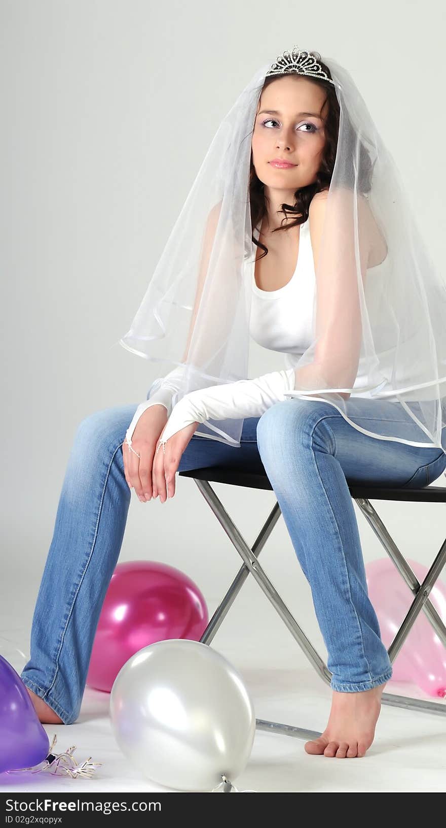 Portrait of the young girl sitting on a chair in jeans and a white vest. The bride in a veil prepares for wedding