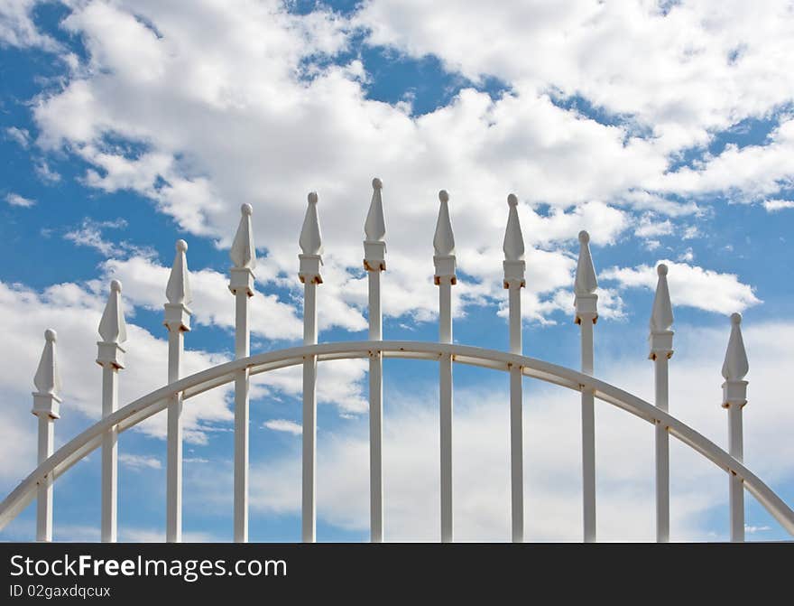 Detail of top of white wrought iron gate against blue sky and clouds