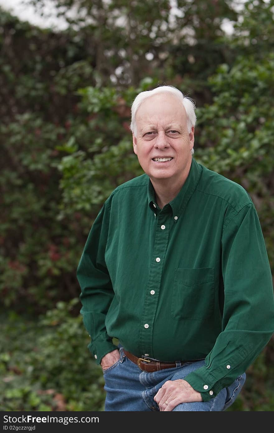 Senior mature man wearing green shirt posing for photograph in front of green bushes.