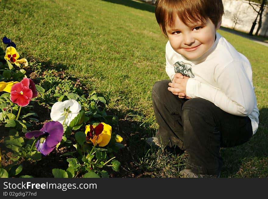 Beautiful smiling boy seating near flowers on green grass, outdoor shot