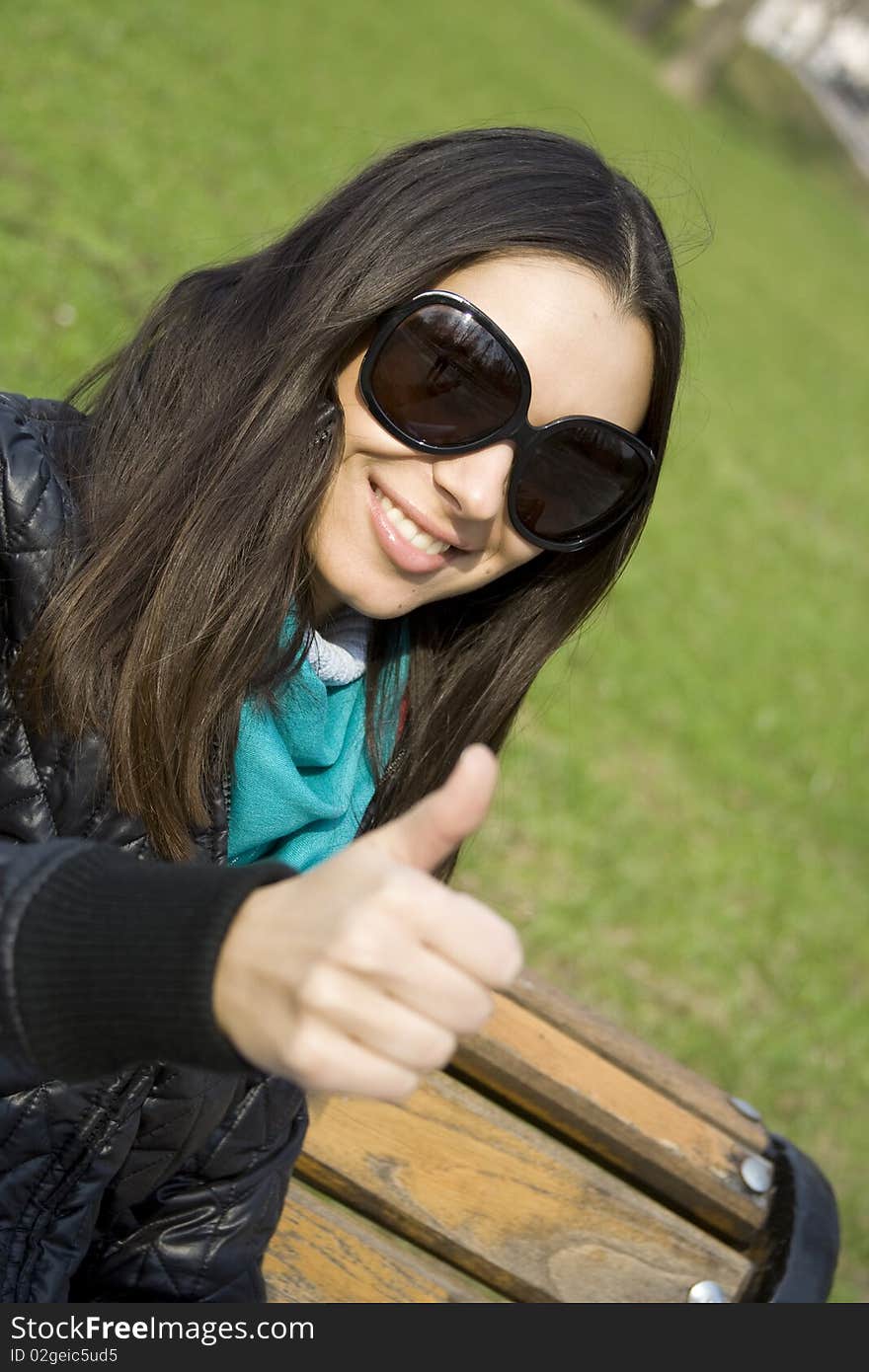 A beautiful young brunette girl in a park sitting on a bench smiling. Thumb sign OK. A beautiful young brunette girl in a park sitting on a bench smiling. Thumb sign OK