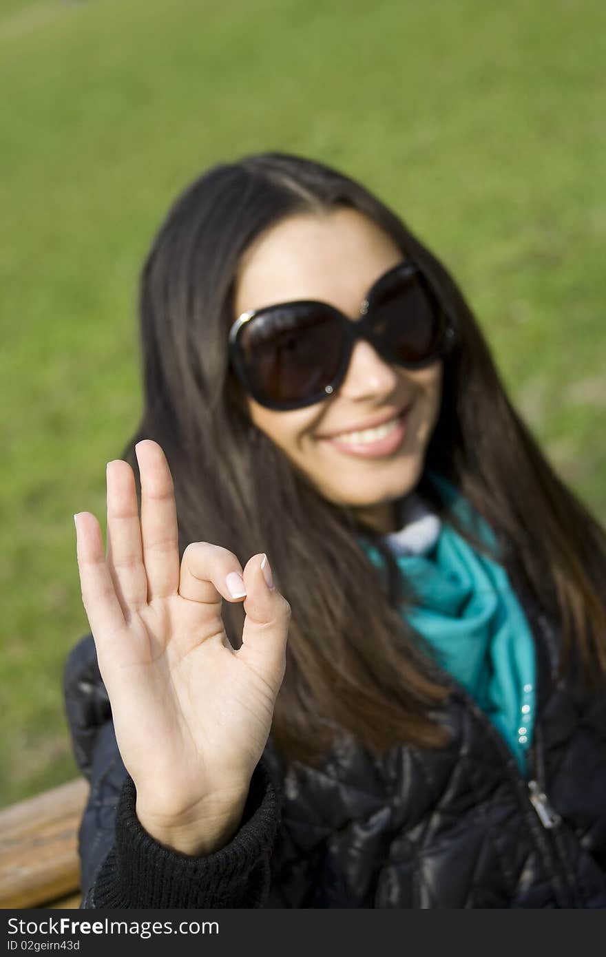 A beautiful young brunette girl in a park sitting on a bench smiling. Hand sign OK. A beautiful young brunette girl in a park sitting on a bench smiling. Hand sign OK