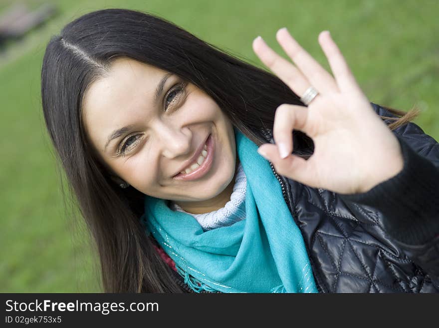 A beautiful young brunette girl in a park sitting on a bench smiling. Hand sign OK