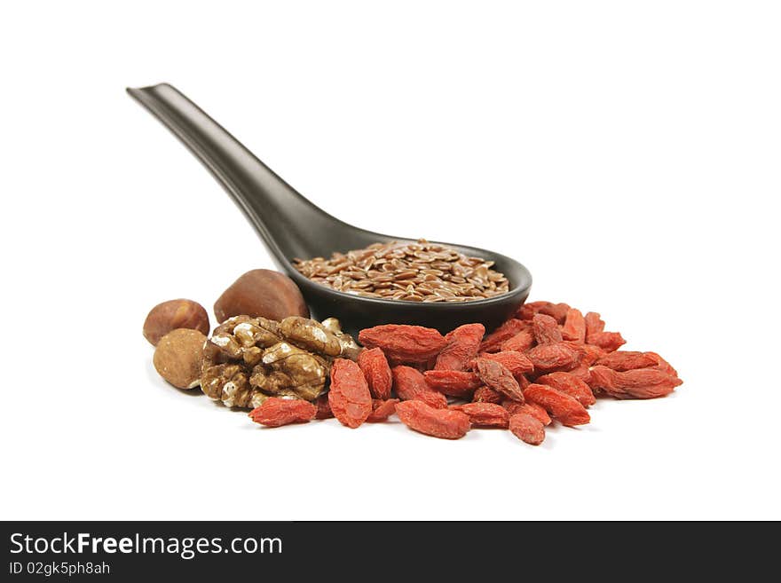 Brown linseed seeds on a small black spoon with mixed nuts and goji berries on a reflective white background. Brown linseed seeds on a small black spoon with mixed nuts and goji berries on a reflective white background