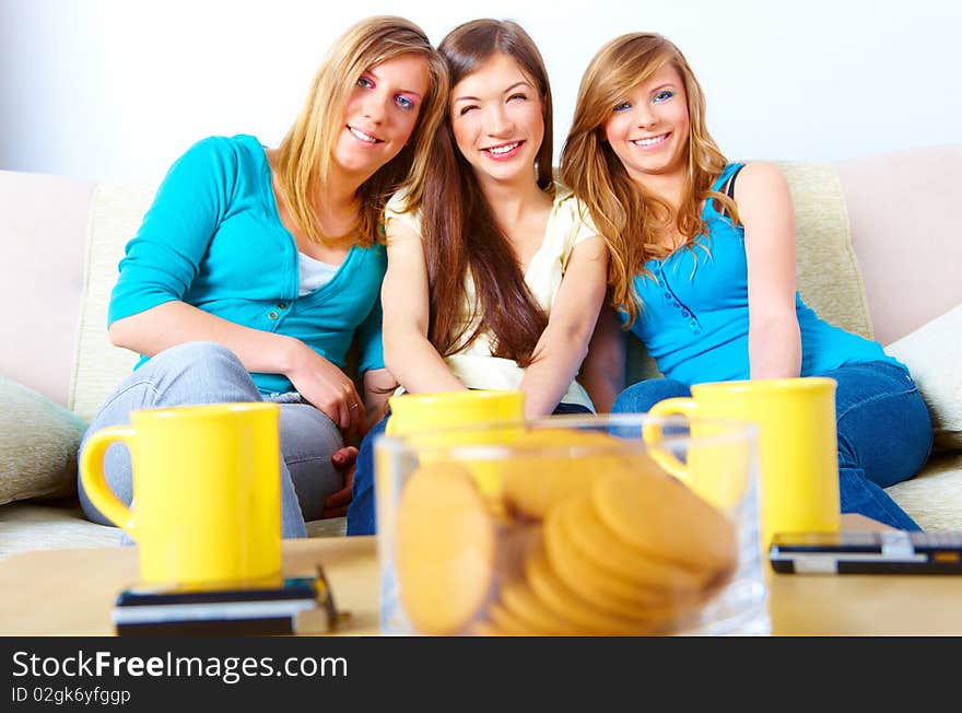 Three happy friends beautiful girls sitting on sofa in home with cups, cookies and phones in the foreground. Three happy friends beautiful girls sitting on sofa in home with cups, cookies and phones in the foreground.