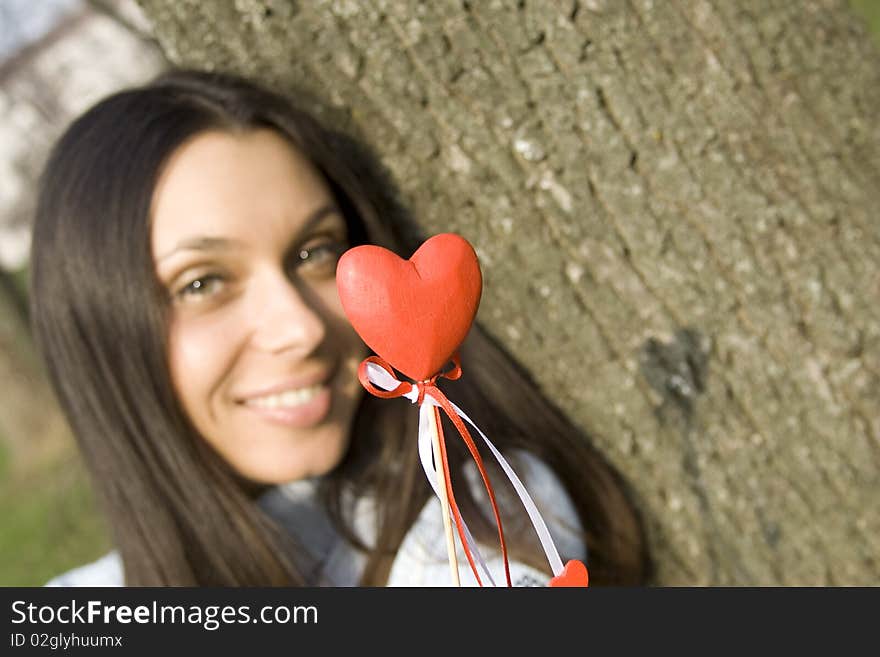 Beautiful girl in a park sitting on the grass near a tree in the hands of a red heart on the wooden stick. Beautiful girl in a park sitting on the grass near a tree in the hands of a red heart on the wooden stick