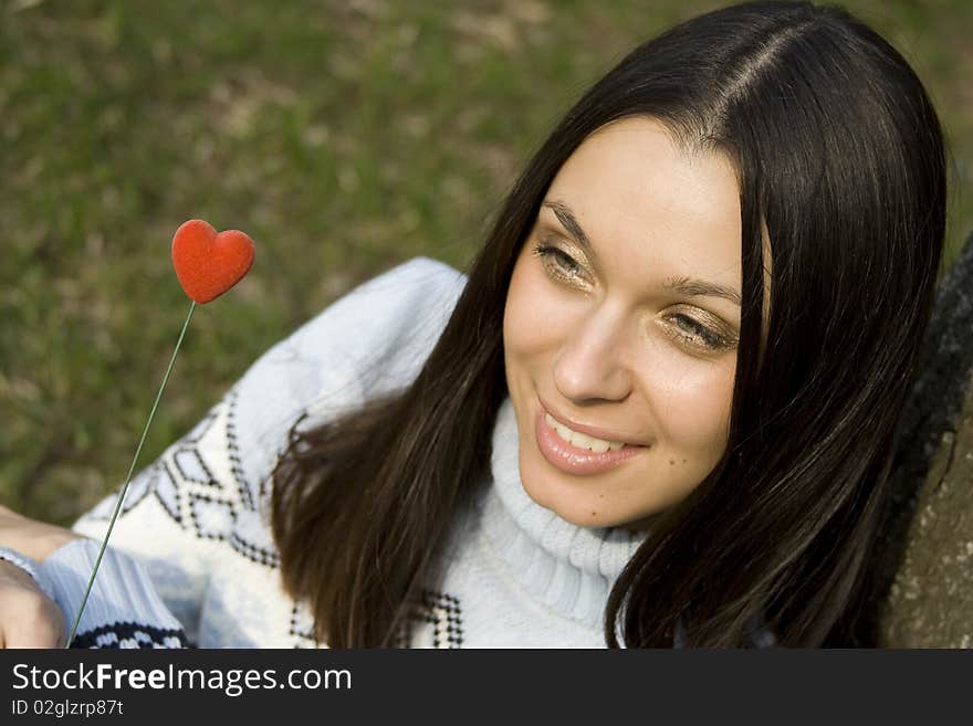 Beautiful girl in a park sitting on the grass near a tree in the hands of a red heart on the wooden stick. Beautiful girl in a park sitting on the grass near a tree in the hands of a red heart on the wooden stick