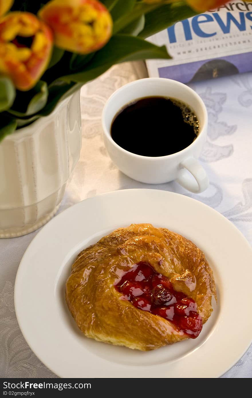 Cherry danish on a white plate with a cup of black coffee, a newspaper and a bunch of tulips. Cherry danish on a white plate with a cup of black coffee, a newspaper and a bunch of tulips