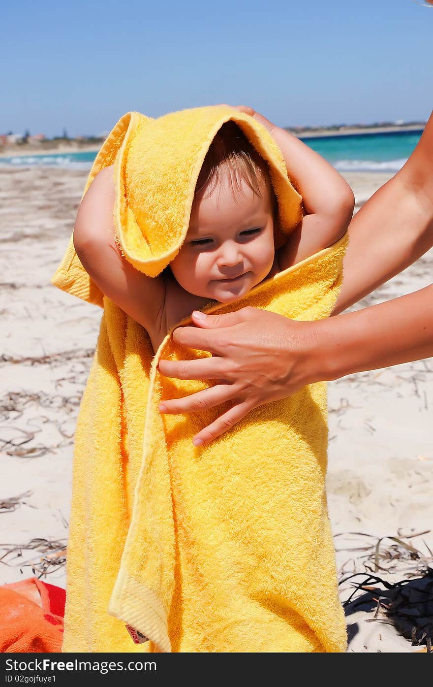 Smiling little girl in a yellow towel on the beach. Caring mother's hands to help her.